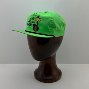 May include: Bright green baseball cap with a black brim and a black rope detail. The cap features the text "Brick Q Town" with a cartoon graphic of a brick chimney and a smiling face.