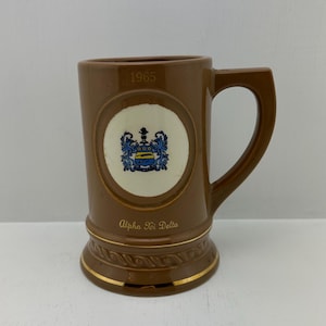 May include: Brown ceramic mug with a handle, featuring a white circle with a blue and gold emblem. The mug has the year "1965" and the text "Alpha Xi Delta" in gold lettering. Gold trim accents the base.