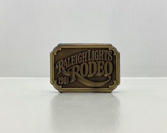 Könnte beinhalten: Rechteckige, bronzefarbene Gürtelschnalle mit den Worten "RALEIGH LIGHTS RODEO 1981" in einem bogenförmigen Design. Die Schnalle hat einen dekorativen Rand und ist vor einem weißen Hintergrund platziert.