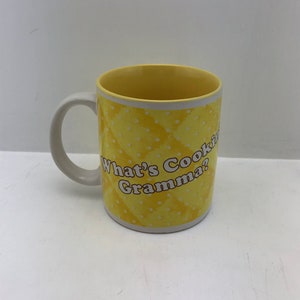 May include: A yellow ceramic mug with a white handle and interior. The mug features a repeating diamond pattern with white polka dots and the text "What's Cookin' Gramma?" in a playful font. The mug is designed for hot beverages.