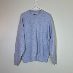 Könnte beinhalten: Ein hellgrauer Pullover mit Rundhalsausschnitt und langen Ärmeln. Der Pullover hat ein kleines weißes Etikett auf der Vorderseite, auf dem "Fruit of the Loom" in schwarzer Schrift steht.