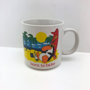 May include: White ceramic mug with a cartoon illustration of a penguin relaxing on a beach with a surfboard. The mug has the words "born to bake" printed on it. The penguin is wearing sunglasses and is next to a bottle.