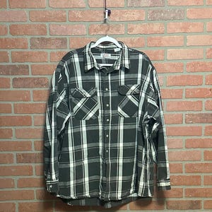 May include: A long-sleeved, button-down shirt with a black, white, and gray plaid pattern. The shirt has two chest pockets and is hanging on a white hanger against a brick wall.