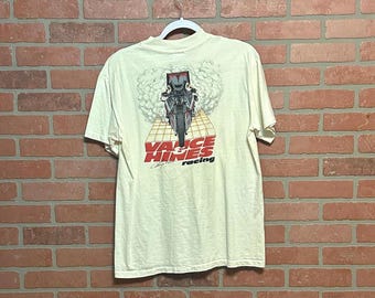 May include: Cream-colored t-shirt featuring a motorcycle graphic with the text "Vance & Hines racing." The graphic shows a motorcycle with a rider. The shirt is hanging against a brick wall.