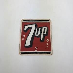 May include: A red and white embroidered patch with the word "7Up" in a white, black, and red font. The patch has a white border and a black outline. There are small white circles around the text.