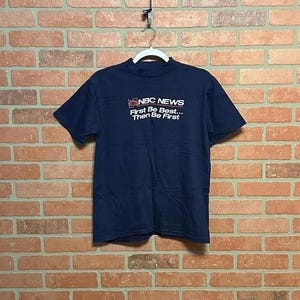 以下が含まれることがあります： NBCニュースのロゴと「First Be Best... Then Be First」の白文字が入ったネイビーブルーのTシャツ。レンガの壁を背景に、白いハンガーにかけられています。
