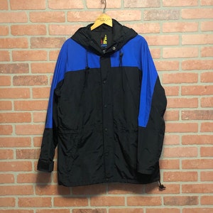 May include: A black and blue hooded jacket hanging on a wooden hanger. The jacket has a black body with blue shoulder and hood panels. It features a button-up front and a drawstring at the bottom. The jacket is hanging against a brick wall.