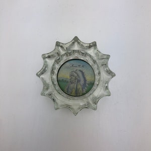 May include: Clear glass ashtray with a starburst design. The centre features a small, circular image of a Native American in a headdress, with the text "Keene, N.H." The ashtray is on a white surface.