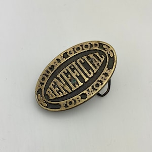 Puede incluir: Hebilla de cinturón ovalada de latón con la palabra "BENEFICIAL" en el centro. Las palabras "YOU'RE A GOOD THING FOR MORE" están alrededor del borde. La hebilla tiene un fondo marrón oscuro y un lazo de metal negro.