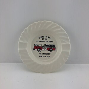 May include: White ceramic plate with a scalloped edge. The plate features a red and white fire truck graphic with the text "ENGINE CO. NO. 1 OF THE PATCHOGUE FIRE DEPT. 95th ANNIVERSARY MARCH 15, 1975".