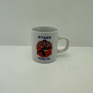 Puede incluir: Taza de cerámica blanca con asa. La taza presenta el texto "NYANG" en azul sobre un gráfico de dibujos animados de un personaje con un atuendo de marinero. Debajo del gráfico está el texto "ROSLYN" en azul. El gráfico está sobre un fondo rojo y naranja.