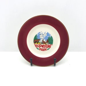 May include: A decorative plate with a maroon rim and a gold edge. The centre features a scene of Yellowstone Park with bears, trees, and a geyser, with the park's name in red and green text. The plate is displayed on a black metal stand.