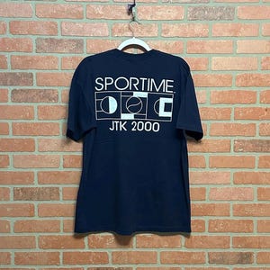 May include: Navy blue t-shirt with the white text "SPORTIME" above a graphic and "JTK 2000". The shirt is hanging on a white hanger against a brick wall.