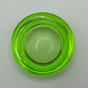 May include: A round, transparent, lime green glass object. The object has a circular shape with a raised, ridged rim. The center of the object is a lighter shade of green. The object is likely a lid or a small container.