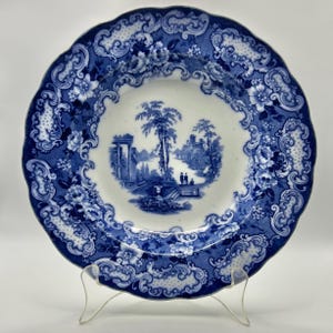 May include: A blue and white ceramic plate with a scalloped edge. The plate features a central landscape scene with trees, figures, and buildings. The border has floral and scrollwork patterns. The plate is displayed on a clear stand.