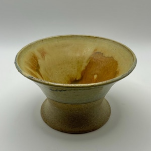May include: A handmade ceramic bowl with a pedestal base. The bowl has a wide, shallow opening and a rounded shape. The interior is a warm, earthy yellow with brown accents, while the exterior features a mix of beige and brown tones.