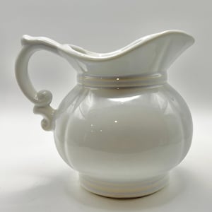 May include: A white ceramic pitcher with a glossy finish, featuring a rounded body, a wide pouring spout, and a curved handle with decorative scrollwork at the base. The pitcher has subtle horizontal bands around the neck and the base.