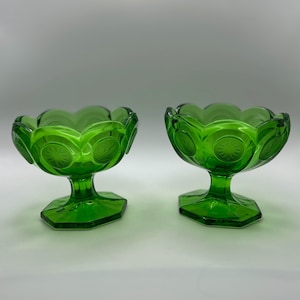 May include: Two green glass dessert bowls with a starburst design. The bowls have a scalloped rim and sit on a pedestal base. The glass is a vibrant, transparent green.