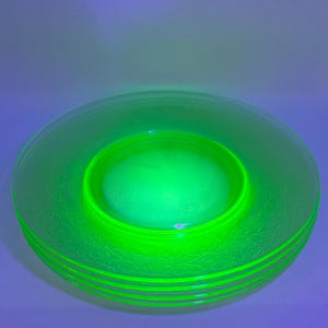 May include: A stack of three round, translucent glass plates that glow with a vibrant, neon green hue under ultraviolet light. Each plate features a smooth, raised rim and a decorative, embossed pattern along the outer edge of the surface.