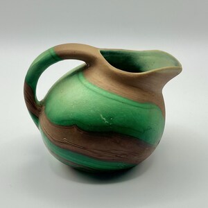 May include: A small, round pitcher with a handle and spout. The pitcher is painted with swirling bands of green and brown. The handle and rim are brown. The pitcher is likely ceramic or pottery.
