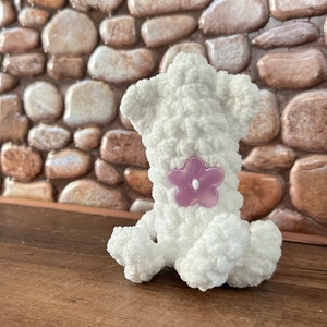 May include: White crocheted stuffed animal shaped like a dog sitting on a wooden surface. The dog has a purple flower sewn on its back.