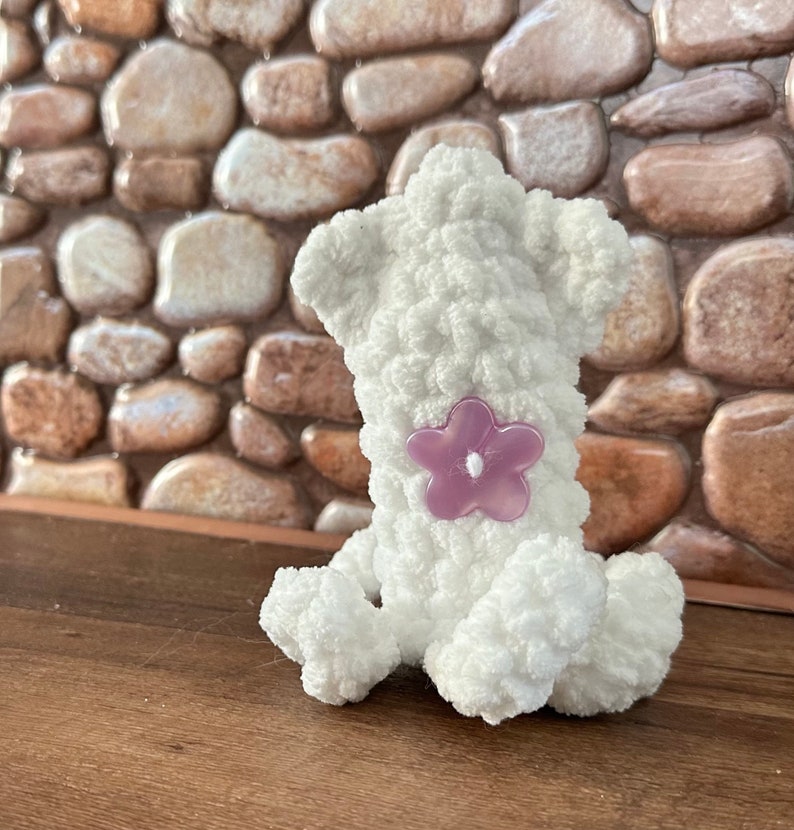 May include: White crocheted stuffed animal shaped like a dog sitting on a wooden surface. The dog has a purple flower sewn on its back.
