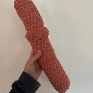 May include: A coral-colored, handmade crochet pillow in a cylindrical shape with a textured surface. The pillow has a thicker, ring-like detail around the middle. The item is held against a plain white background.