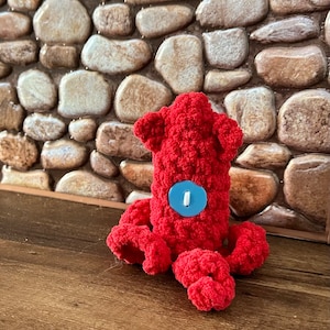 May include: A red crocheted octopus with a blue button for an eye. The octopus is sitting on a brown textured surface.