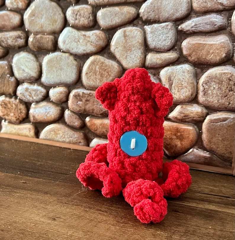 May include: A red crocheted octopus with a blue button for an eye. The octopus is sitting on a brown textured surface.