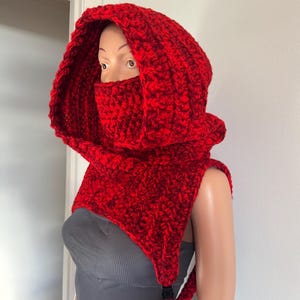 May include: A handmade, red crochet hood with a matching face covering. The hood is designed to cover the head and neck, with a built-in face mask. The hood has a textured, chunky knit appearance and a black buckle closure.