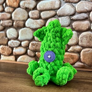May include: A green crocheted octopus with a purple button on its body. The octopus is sitting on a wooden surface.