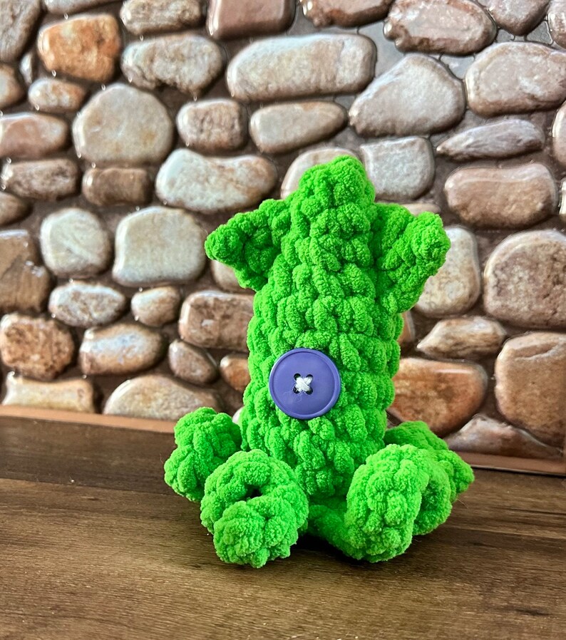 May include: A green crocheted octopus with a purple button on its body. The octopus is sitting on a wooden surface.