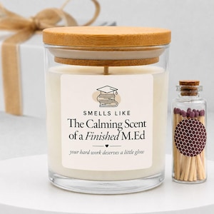 May include: A clear glass candle jar with a light wood lid and a white label featuring a graduation cap icon, the text "SMELLS LIKE The Calming Scent of a Finished M.Ed" and "your hard work deserves a little glow," next to a small glass match jar.