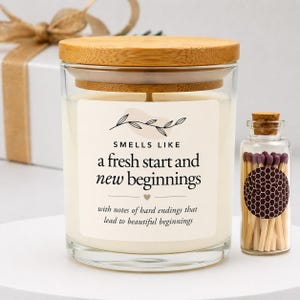 May include: A clear glass candle jar with a light-coloured wax, a bamboo lid, and a label reading "SMELLS LIKE a fresh start and new beginnings with notes of hard endings that lead to beautiful beginnings." Next to it is a small glass bottle of matches.