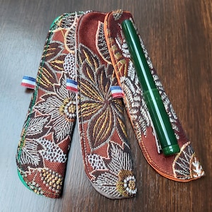 Op de afbeelding: Drie pennenzakken met bloempatronen in bruin, goud en wit. Elke etui heeft een klein lint met rood-wit-blauwe strepen. Eén etui bevat een groene pen. De etuis liggen op een houten oppervlak.