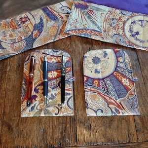 Op de afbeelding: Drie pennen worden getoond in een pennenhouder met een kleurrijk, gepatroneerd ontwerp. De pennenhouder is gemaakt van stof en heeft een blauw, rood en gouden kleurenschema. De pennen zijn zwart, blauw en bruin.