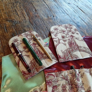 May include: Three pens are displayed in a fabric pen case with a floral design. The case is beige with a red and green floral pattern. The pens are black, green, and gold.