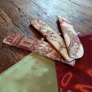 Op de afbeelding: Drie met stof beklede bladwijzers met bloemen- en geometrische patronen. De bladwijzers zijn rood, wit en blauw met een rood, wit en blauwe rand. De bladwijzers liggen op een groene en rode stoffen ondergrond.