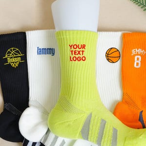 Op de afbeelding: Een verzameling basketbalsokken in verschillende kleuren, waaronder zwart, wit, geel en oranje. Elke sok heeft een uniek ontwerp, zoals een basketbalafbeelding, gepersonaliseerde tekst en spelersnummers. De sokken worden getoond op een witte mannequinbeen.