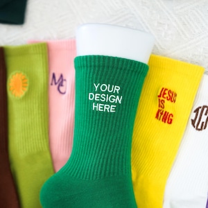 Peut inclure: Un ensemble de neuf chaussettes colorées avec différents motifs brodés. Les motifs comprennent un tournesol, les lettres "MC", les mots "Your Design Here", les mots "Jesus is King", les lettres "JCF", et un autre tournesol.