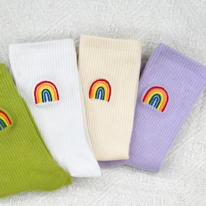 Puede incluir: Un conjunto de cuatro calcetines acanalados en verde lima, blanco, crema y lavanda, cada uno con un pequeño motivo de arcoíris bordado con rayas rojas, naranjas, amarillas, verdes y azules en la parte superior de la tela.