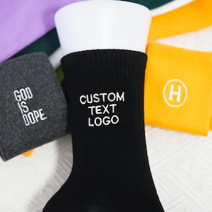 May include: Black ribbed socks with white embroidered text reading "CUSTOM TEXT LOGO." A gray sock with "GOD IS DOPE" embroidered in white is also visible. An orange sock with a white "H" logo is also visible.