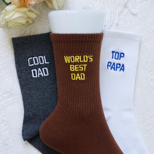 May include: Three pairs of crew socks with text embroidered on them. One pair is dark gray with "COOL DAD" in white. The second pair is brown with "WORLD'S BEST DAD" in yellow. The third pair is white with "TOP PAPA" in blue.