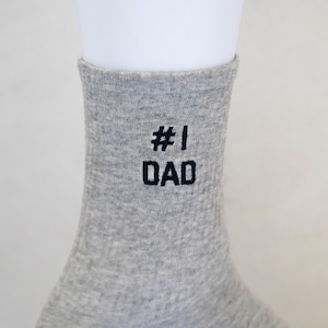 May include: A gray sock with the text "#1 DAD" embroidered in black. The sock is made of a soft, ribbed material and is displayed on a white mannequin foot. The sock is a gift for Father's Day.