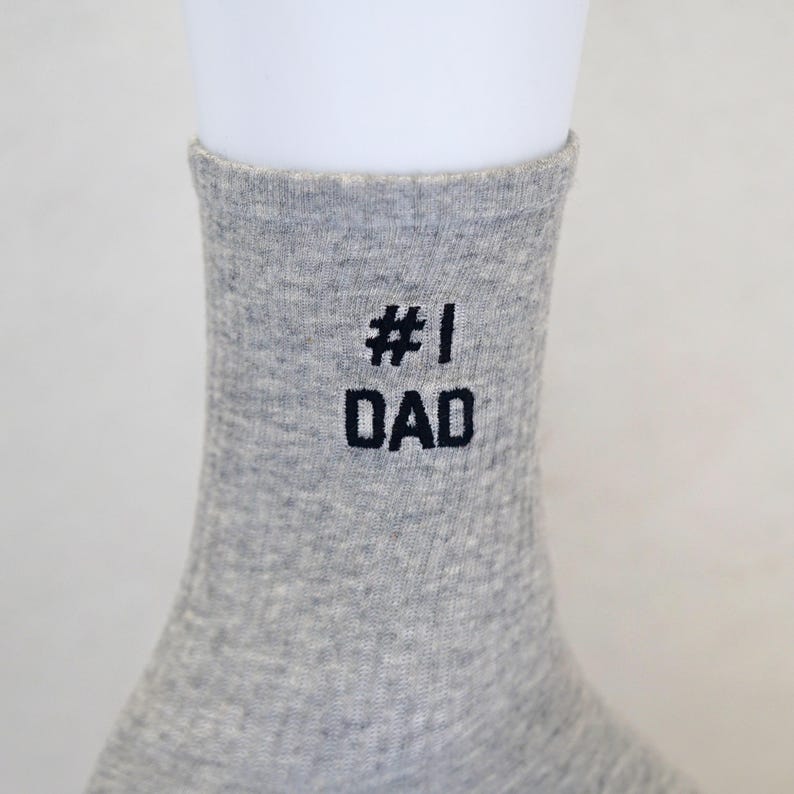 May include: A gray sock with the text "#1 DAD" embroidered in black. The sock is made of a soft, ribbed material and is displayed on a white mannequin foot. The sock is a gift for Father's Day.