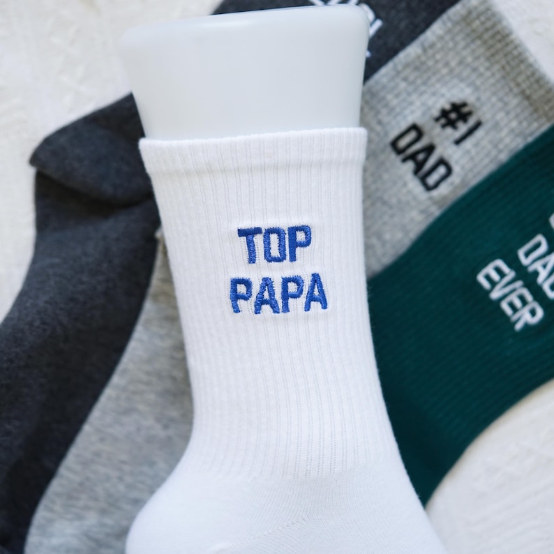 May include: White sock with "TOP PAPA" embroidered in blue. Other socks in the background include gray and dark green socks with "#1 DAD" and "DAD EVER" in white. The socks are on a white surface.