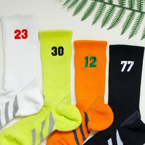 Puede incluir: Cuatro calcetines deportivos en blanco, verde neón, naranja y negro, cada uno con un número diferente: 23, 30, 12 y 77. Los calcetines tienen una textura acanalada y detalles grises en los talones y las suelas. Una hoja de helecho en el fondo.