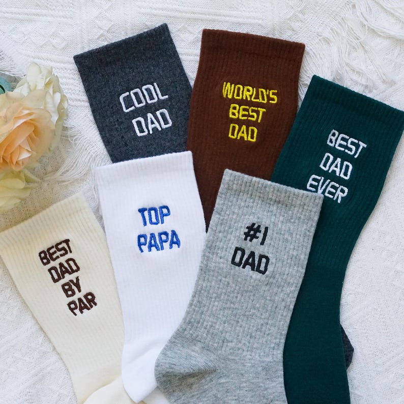 May include: A collection of colorful socks with text embroidered on them. The socks are in various colors, including gray, white, brown, and green. The text on the socks includes phrases like "COOL DAD", "WORLD'S BEST DAD", and "BEST DAD EVER".