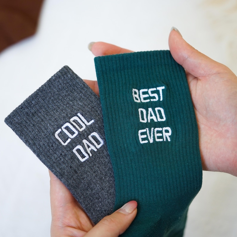 May include: Two socks are held in a person's hands. One sock is dark gray with the words "COOL DAD" in white. The other sock is dark green with the words "BEST DAD EVER" in white. The socks are made of a ribbed knit material.