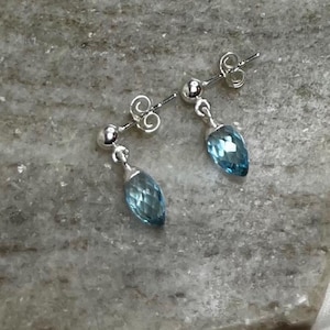 May include: A pair of silver stud earrings with teardrop-shaped, faceted blue gemstones. The earrings have a post and ball design, with a small, decorative swirl detail above the post. The gemstones are a light, translucent blue.